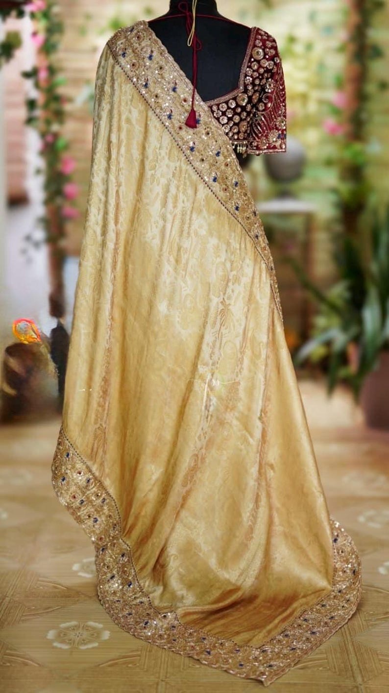 May include: A gold-colored sari with intricate floral patterns and a decorative border. The sari is draped over a mannequin, revealing a maroon blouse with detailed embellishments. The border is adorned with sequins and blue accents.