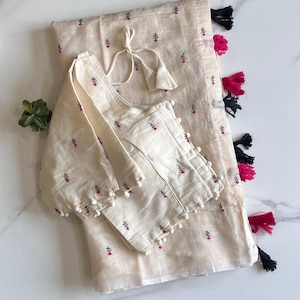 May include: Cream-colored embroidered blouse and matching fabric with a floral pattern. The blouse has long sleeves with pom-pom trim and a tie closure. The fabric has tassels in black and fuchsia.