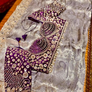 May include: A luxurious, ornate purple and gold embroidered bodice and sleeves, with intricate beadwork and sequin details. The garment is set on a silver-gray fabric with a subtle pattern, suggesting formal wear or a special occasion ensemble.