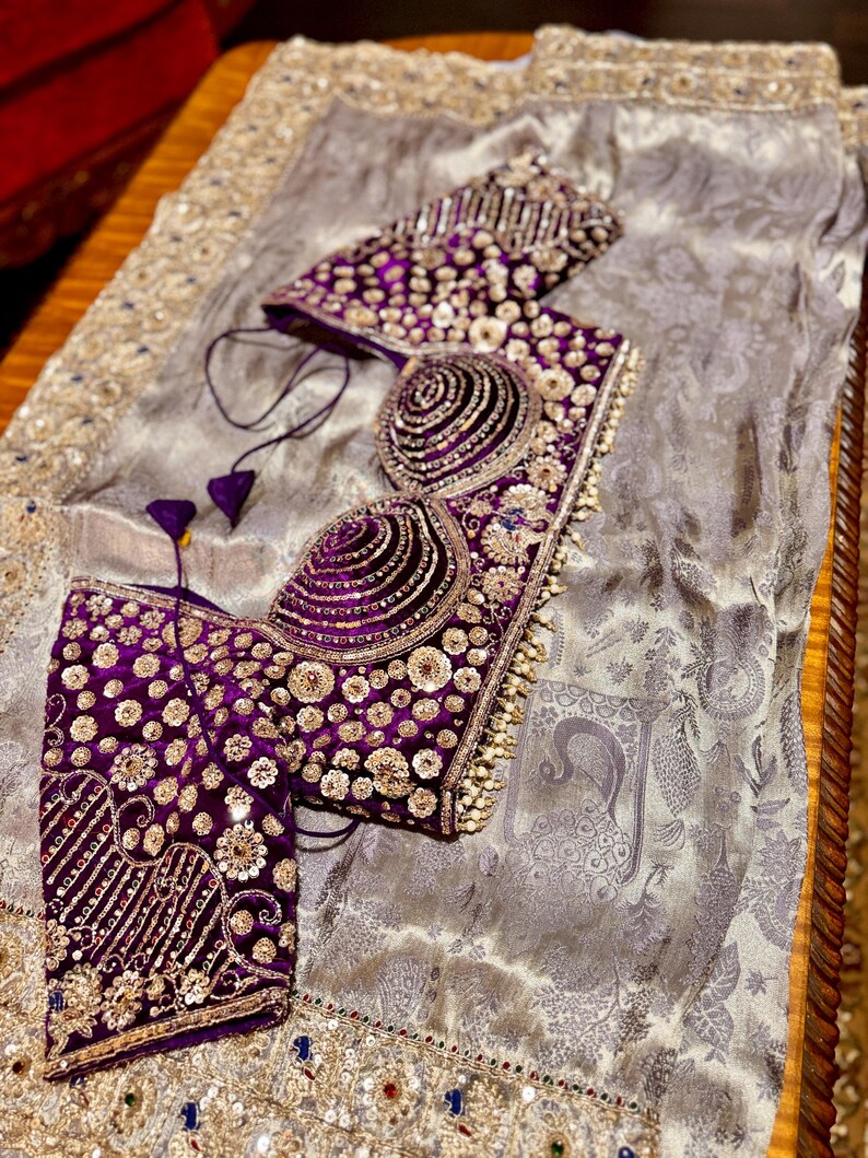 May include: A luxurious, ornate purple and gold embroidered bodice and sleeves, with intricate beadwork and sequin details. The garment is set on a silver-gray fabric with a subtle pattern, suggesting formal wear or a special occasion ensemble.