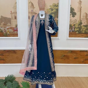 May include: A teal-colored formal dress with intricate gold embroidery and embellishments. The dress features a matching jacket with similar detailing and a sheer, light pink scarf. The outfit is displayed on a mannequin.