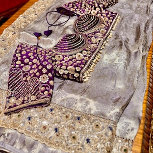May include: A luxurious ensemble featuring a silver-gray fabric with an ornate, embroidered border. The set includes a richly embellished, deep purple bodice and matching accessories, adorned with gold and pearl-like accents. The intricate design suggests formal wear.