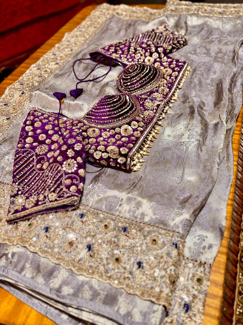 May include: A luxurious ensemble featuring a silver-gray fabric with an ornate, embroidered border. The set includes a richly embellished, deep purple bodice and matching accessories, adorned with gold and pearl-like accents. The intricate design suggests formal wear.
