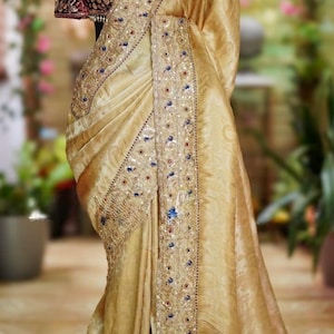 May include: A gold-colored sari with intricate embroidery and embellishments. The sari is draped over a mannequin, showcasing its flowing fabric and detailed design. The blouse is a deep burgundy with ornate beadwork.