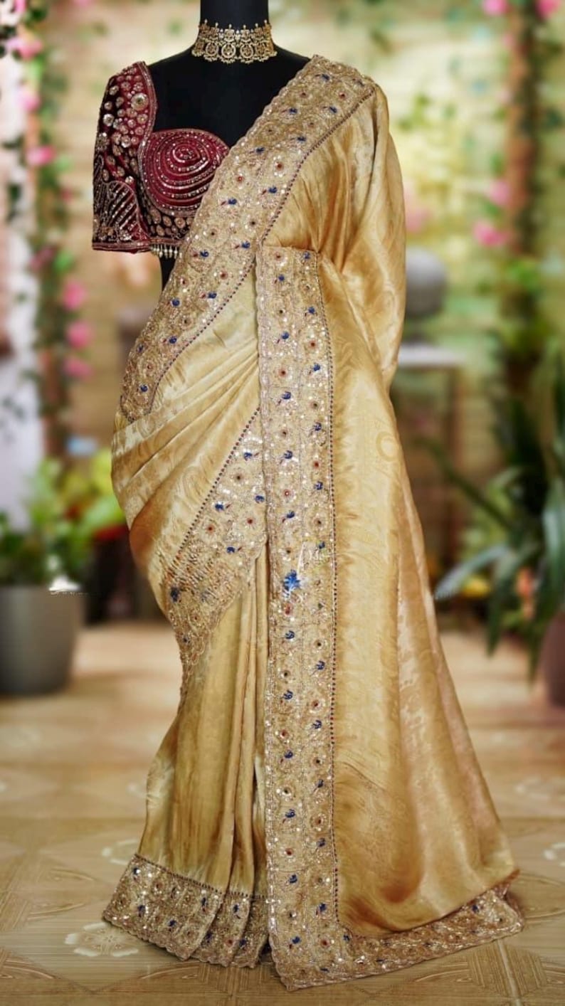 May include: A gold-colored sari with intricate embroidery and embellishments. The sari is draped over a mannequin, showcasing its flowing fabric and detailed design. The blouse is a deep burgundy with ornate beadwork.