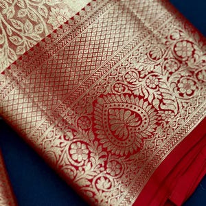 May include: Close-up of a red and gold textile with intricate floral and geometric patterns. The fabric features a detailed border and a contrasting cream-colored section with a leafy design. The textile is folded, revealing its layered construction.