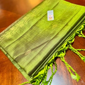 May include: Folded green fabric with a textured surface and a decorative border. The fabric has a fringe of twisted green threads with tassels. A small tag is attached to the fabric.