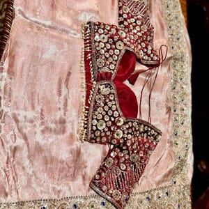 May include: A traditional Indian garment set featuring a burgundy velvet blouse with intricate beadwork and embellishments. The blouse is adorned with sparkling sequins and pearls. It rests on a light pink, patterned fabric with a decorative border.
