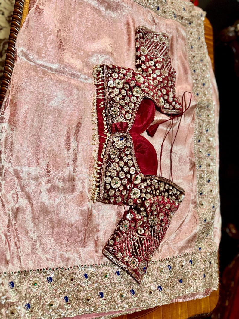May include: A traditional Indian garment set featuring a burgundy velvet blouse with intricate beadwork and embellishments. The blouse is adorned with sparkling sequins and pearls. It rests on a light pink, patterned fabric with a decorative border.
