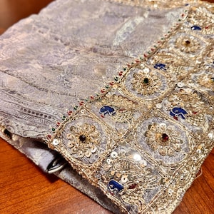 May include: A folded, ornate textile with a silver-gray paisley pattern. The edge is embellished with gold embroidery, sequins, and colorful gemstones. The fabric appears soft and luxurious, with intricate detailing.