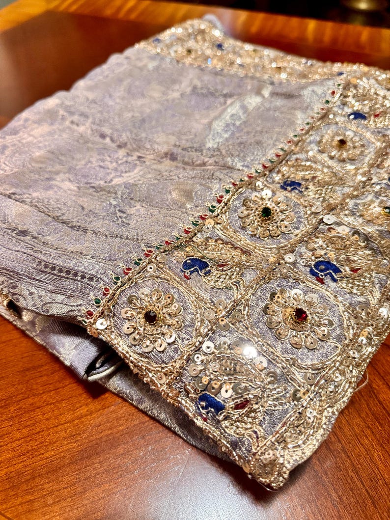 May include: A folded, ornate textile with a silver-gray paisley pattern. The edge is embellished with gold embroidery, sequins, and colorful gemstones. The fabric appears soft and luxurious, with intricate detailing.