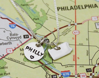 May include: Two silver necklaces with charms. One charm is a round disc with the word "PHILLY" and a heart. The other charm is a silver outline of the state of Pennsylvania. The necklaces are laying on a map of Philadelphia.