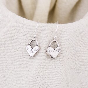 May include: A pair of silver heart-shaped dangle earrings. Each earring features a textured heart pendant suspended from a simple hook. The earrings are displayed on a cream-colored fabric, creating a soft, neutral backdrop.