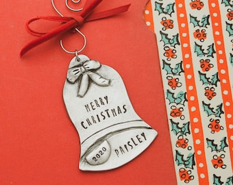 Pode incluir: Um enfeite de sino de prata com uma fita e um laço vermelhos. O sino tem gravadas as palavras "Merry Christmas" e "Paisley", juntamente com o ano "2020".