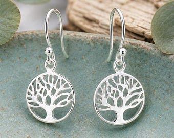 May include: A pair of silver drop earrings featuring a circular pendant with a cut-out tree of life design. Each earring hangs from a curved silver hook, showcasing a polished metallic finish and intricate branching details within the round frame.