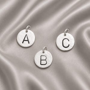 May include: Three circular silver-toned metal charms, each featuring a single stamped letter A, B, or C in black. The surface of each charm has a hammered, textured finish and includes a small jump ring at the top for attachment to jewelry.