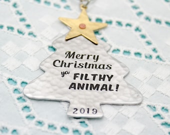 Op de afbeelding: Zilverkleurige kerstboomversiering met de tekst "Merry Christmas ya FILTHY ANIMAL!" en het jaar 2019. Het heeft een gehamerde textuur en een gouden ster aan de bovenkant.
