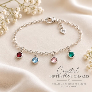 May include: A silver-toned chain bracelet featuring four round bezel-set crystal charms in red, light blue, pink, and green. The bracelet includes a small heart-shaped charm near the lobster clasp. Text reads: Crystal BIRTHSTONE CHARMS.
