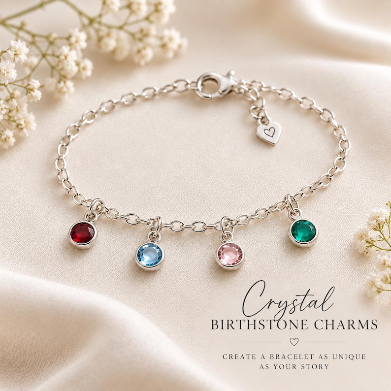 May include: A silver-toned chain bracelet featuring four round bezel-set crystal charms in red, light blue, pink, and green. The bracelet includes a small heart-shaped charm near the lobster clasp. Text reads: Crystal BIRTHSTONE CHARMS.