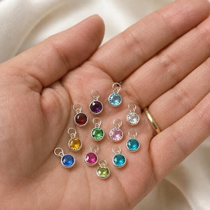 May include: A collection of thirteen small, round, silver-toned charms, each featuring a faceted gemstone in a variety of vibrant colors including blue, purple, red, yellow, green, pink, and clear, all equipped with a small circular jump ring.