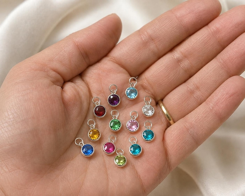 May include: A collection of thirteen small, round, silver-toned charms, each featuring a faceted gemstone in a variety of vibrant colors including blue, purple, red, yellow, green, pink, and clear, all equipped with a small circular jump ring.