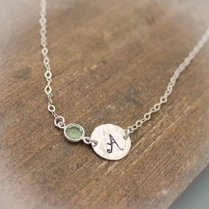 Puede incluir: Un collar de plata con una cadena delicada. Presenta una pequeña gema verde redonda y un disco de plata martillada con la letra "A" grabada. El collar se muestra sobre una superficie de madera.
