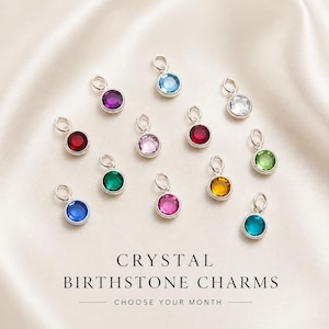 May include: A collection of twelve circular silver charms, each featuring a different colored crystal birthstone. The charms are arranged in a scattered pattern. Below the charms, the text reads: CRYSTAL BIRTHSTONE CHARMS CHOOSE YOUR MONTH.