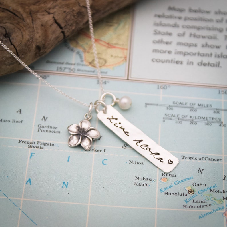 May include: Silver necklace with a small flower charm, a silver bar charm with the text "Live Aloha" and a small pearl charm. The necklace is photographed on a map of the Hawaiian islands.