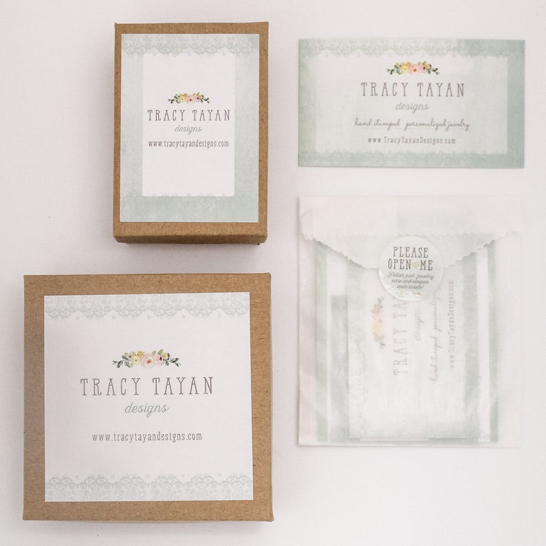 以下が含まれることがあります： Tracy Tayan Designsのパッケージの俯瞰図。画像には、2つの小さな茶色の箱、名刺、小さな紙袋が含まれています。各アイテムには、Tracy Tayan Designsのロゴとウェブサイトアドレスが表示されています。名刺には「手作りのパーソナライズされたジュエリー」と書かれています。
