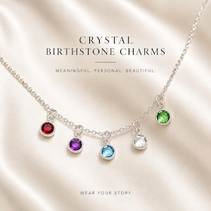 May include: A silver chain necklace featuring five dangling round crystal birthstone charms in red, purple, light blue, clear, and green. The text CRYSTAL BIRTHSTONE CHARMS, MEANINGFUL. PERSONAL. BEAUTIFUL. and WEAR YOUR STORY is printed on the image.