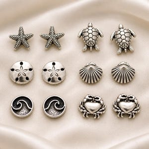 Puede incluir: Una colección de doce pendientes de botón de metal plateado dispuestos en pares. El conjunto incluye estrellas de mar, tortugas marinas, dólares de arena, conchas, olas del mar y cangrejos. Cada pieza presenta texturas detalladas.