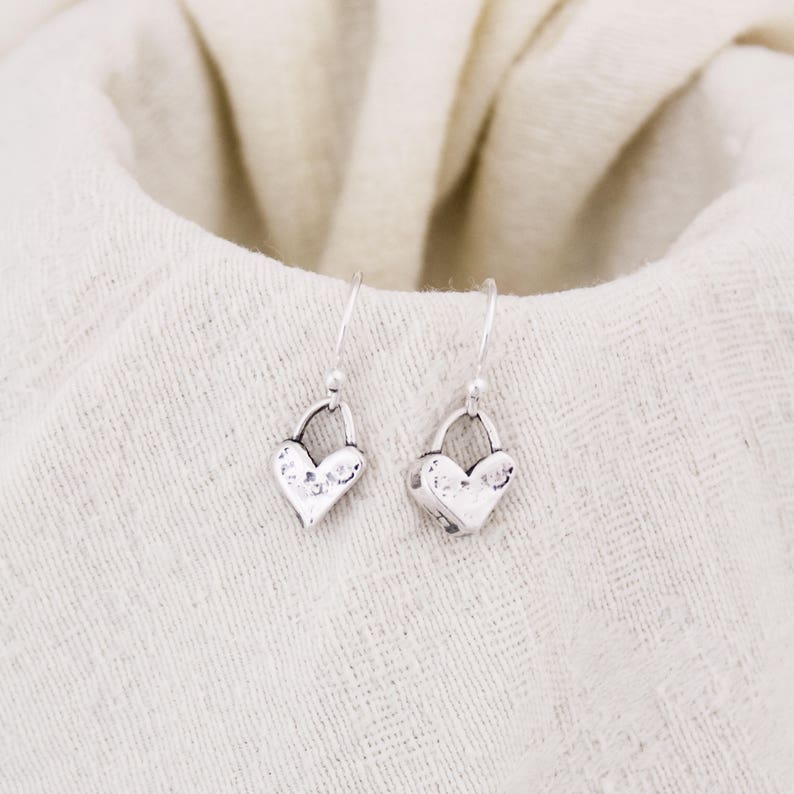 May include: A pair of silver heart-shaped dangle earrings. Each earring features a textured heart pendant suspended from a simple hook. The earrings are displayed on a cream-colored fabric, creating a soft, neutral backdrop.