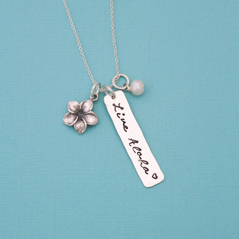 May include: Silver necklace with a small flower charm, a pearl charm, and a rectangular tag that says "Live Aloha" with a heart symbol.