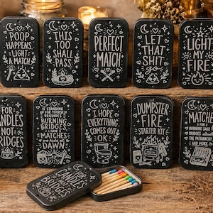 May include: A collection of rectangular black metal match tins featuring white hand-lettered text and illustrations. Each tin displays unique phrases like Poop Happens, This Too Shall Pass, Perfect Match, Let That Shit Go, and Light My Fire.