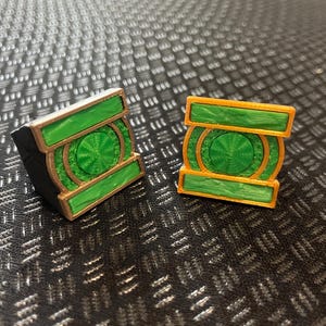 Puede incluir: Dos emblemas de forma cuadrada con un diseño circular verde central rodeado por un borde de color dorado. Los emblemas tienen una apariencia texturizada y en capas con un centro verde prominente y barras horizontales en la parte superior e inferior, mostrando una estructura geométrica distinta.