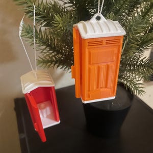 May include: Two portable toilet ornaments hanging from a tree. One is bright orange with a white roof and base, while the other is red with a white roof and base, featuring an open door that reveals a small white toilet seat inside each unit.