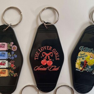 K&ouml;nnte beinhalten: Drei schwarze Schl&uuml;sselanh&auml;nger in Form von Motelzimmerschl&uuml;sseln. Die Schl&uuml;sselanh&auml;nger haben verschiedene Designs: einer mit einem Muster aus Diet Coke-Dosen und Blumen, einer mit dem Text "The Lover Girls Social Club" und zwei roten Kirschen und einer mit dem Text "I Beg You Don't Embarrass Me" und einer Comic-Frau in einem gelben Kleid.