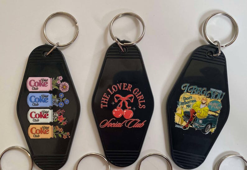 K&ouml;nnte beinhalten: Drei schwarze Schl&uuml;sselanh&auml;nger in Form von Motelzimmerschl&uuml;sseln. Die Schl&uuml;sselanh&auml;nger haben verschiedene Designs: einer mit einem Muster aus Diet Coke-Dosen und Blumen, einer mit dem Text "The Lover Girls Social Club" und zwei roten Kirschen und einer mit dem Text "I Beg You Don't Embarrass Me" und einer Comic-Frau in einem gelben Kleid.