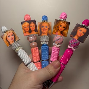 May include: Five pens featuring square toppers with printed images of doll faces with various expressions. Each pen has a textured, glittery barrel in white, orange, blue, light pink, or dark pink, accented with decorative beads and rhinestones.