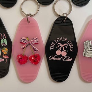 K&ouml;nnte beinhalten: Vier Schl&uuml;sselanh&auml;nger mit verschiedenen Designs. Der erste Schl&uuml;sselanh&auml;nger ist schwarz mit dem Wort "FLORIDA" und Bildern eines Flamingos, Palmen und eines Cocktails. Der zweite Schl&uuml;sselanh&auml;nger ist pink mit einem Paar Kirschen mit einer Schleife oben drauf. Der dritte Schl&uuml;sselanh&auml;nger ist schwarz mit dem Text "THE LOVER GIRLS SOCIAL CLUB". Der vierte Schl&uuml;sselanh&auml;nger ist pink mit einem Zeitungsartikel, auf dem "Breaking Heart" steht.