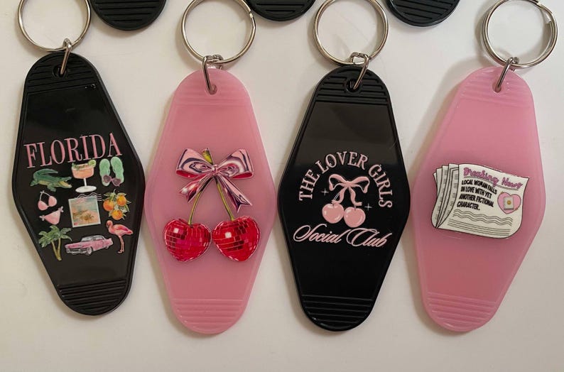 K&ouml;nnte beinhalten: Vier Schl&uuml;sselanh&auml;nger mit verschiedenen Designs. Der erste Schl&uuml;sselanh&auml;nger ist schwarz mit dem Wort "FLORIDA" und Bildern eines Flamingos, Palmen und eines Cocktails. Der zweite Schl&uuml;sselanh&auml;nger ist pink mit einem Paar Kirschen mit einer Schleife oben drauf. Der dritte Schl&uuml;sselanh&auml;nger ist schwarz mit dem Text "THE LOVER GIRLS SOCIAL CLUB". Der vierte Schl&uuml;sselanh&auml;nger ist pink mit einem Zeitungsartikel, auf dem "Breaking Heart" steht.