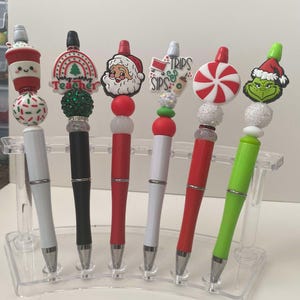 Puede incluir: Seis bolígrafos con temática navideña con diferentes adornos coloridos, incluyendo un muñeco de nieve, un bastón de caramelo, un Grinch y un Papá Noel. Los bolígrafos se muestran en un soporte de plástico transparente.