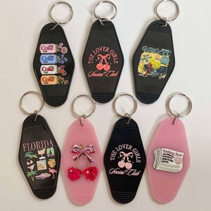 K&ouml;nnte beinhalten: Sieben Vintage-Motel-Schl&uuml;sselanh&auml;nger mit verschiedenen Designs. Die Schl&uuml;sselanh&auml;nger sind schwarz und rosa mit verschiedenen Bildern, darunter ein Coca-Cola-Design, Kirschen, eine Zeitung und ein Flamingo. Die Schl&uuml;sselanh&auml;nger tragen den Text "The Lover Girls Social Club" und "I Beg You Don't Embarrass Me".