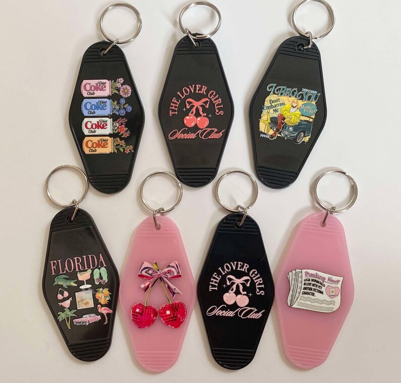 K&ouml;nnte beinhalten: Sieben Vintage-Motel-Schl&uuml;sselanh&auml;nger mit verschiedenen Designs. Die Schl&uuml;sselanh&auml;nger sind schwarz und rosa mit verschiedenen Bildern, darunter ein Coca-Cola-Design, Kirschen, eine Zeitung und ein Flamingo. Die Schl&uuml;sselanh&auml;nger tragen den Text "The Lover Girls Social Club" und "I Beg You Don't Embarrass Me".