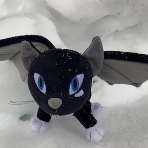 May include: A black plush cat with gray bat wings, blue eyes, and white paws rests on a snowy surface. The cat has a gray nose and whiskers. The wings are gray with black trim. The cat is a whimsical, fantasy-themed item.