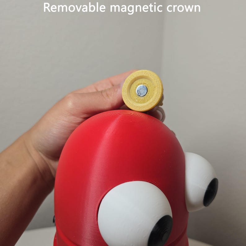 May include: A red, rounded figure with large white eyes featuring black pupils, topped with a small, yellow, circular, removable magnetic crown. The text "Removable magnetic crown" is visible at the top of the image, describing the accessory.