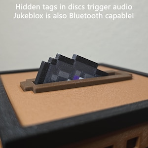 May include: A square Jukeblox device with a tan top surface and black frame. A pixelated black and purple disc is inserted into a rectangular slot. Text reads: Hidden tags in discs trigger audio. Jukeblox is also Bluetooth capable!