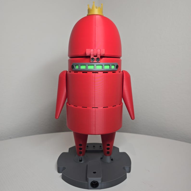 May include: A red robot figurine with a gold crown on its head, a horizontal green light strip across the chest, and two arms extending from the sides. The robot stands on a circular gray base with a small black circular port at the front center.