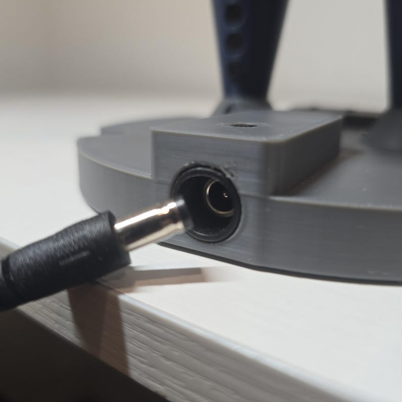 May include: A gray 3D-printed circular base featuring a central power input port. A black cylindrical power cable connector is positioned directly in front of the port, ready to be inserted into the circular opening of the device base.