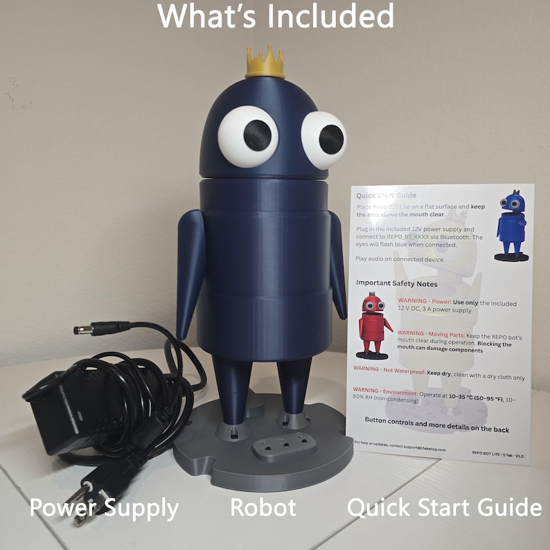 May include: A dark blue robot figure with large white eyes and a small gold crown on its head, standing on a gray base. Included are a black power supply cord with a plug and a white Quick Start Guide document with printed safety instructions.